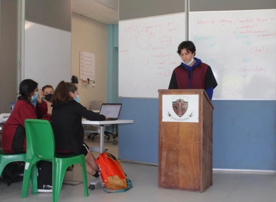 A student speaking from a podium in a classroom round while three others judge and flow at a table, with the case mapped out on the whiteboard behind